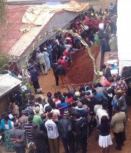 A crowd of mourners outside the family's home with the burial site adjacent to the mount of dirt.